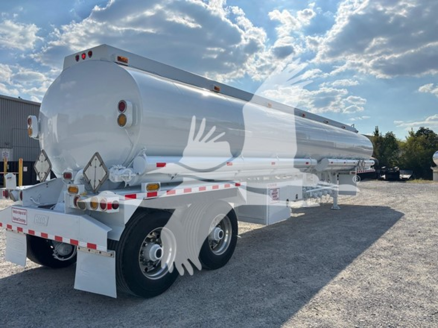 Semi-trailer tanker in a yard with blue sky and clouds; white cylindrical tank on dual rear axles, rear lights and safety reflectors visible.