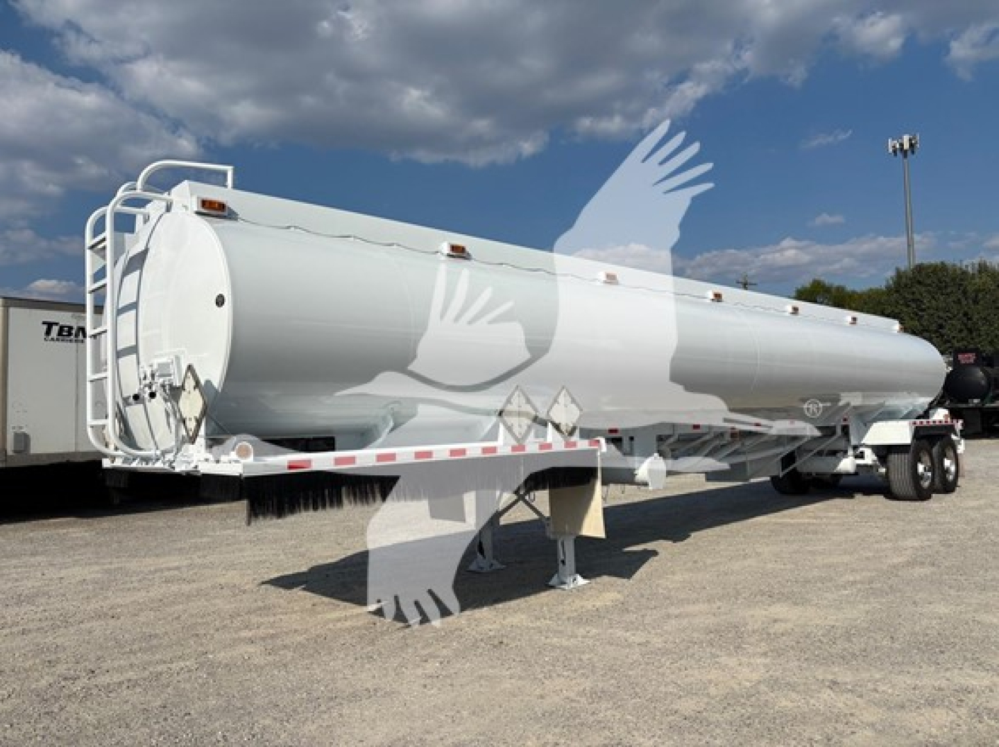 Large white Heil DOT406 tanker trailer on a gravel lot, cylindrical tank with ladder at rear and landing gear visible.
