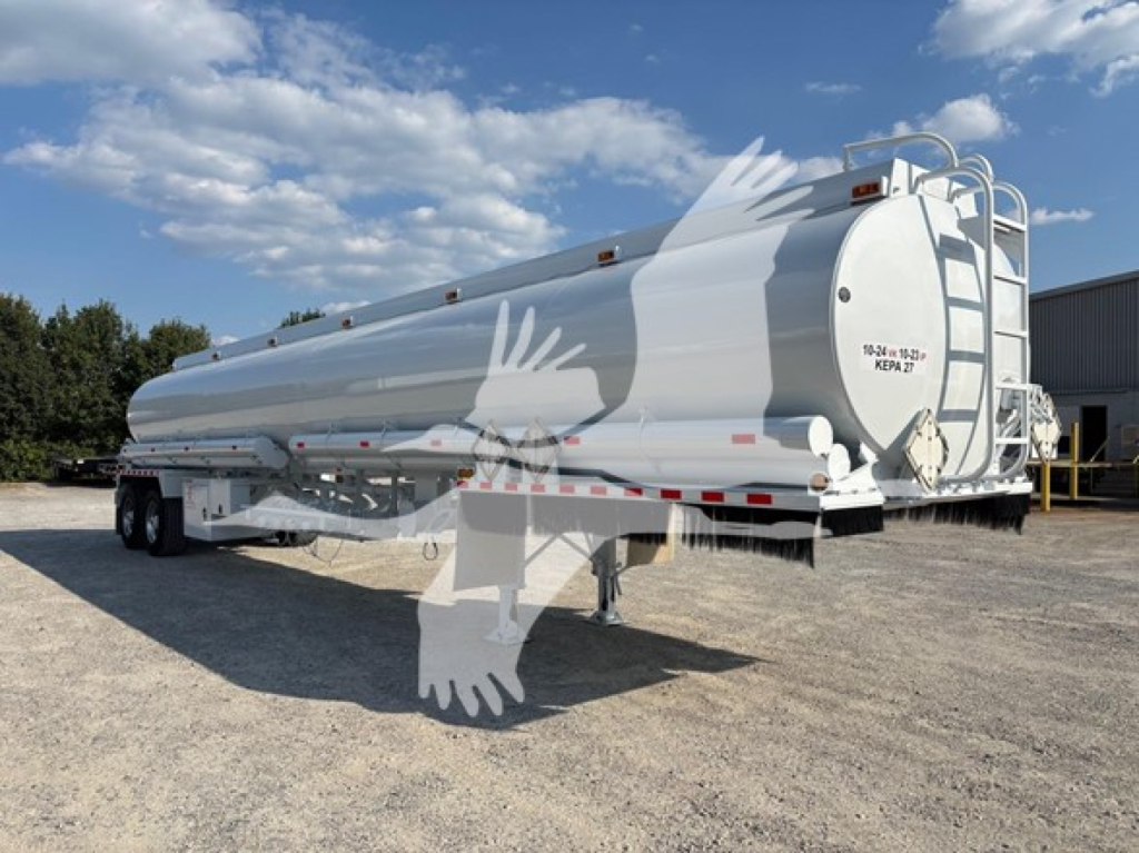 Trailer in a yard showing a white Heil DOT406 tanker with tandem axles and rear access ladder, side-mounted discharge, and warning decals.