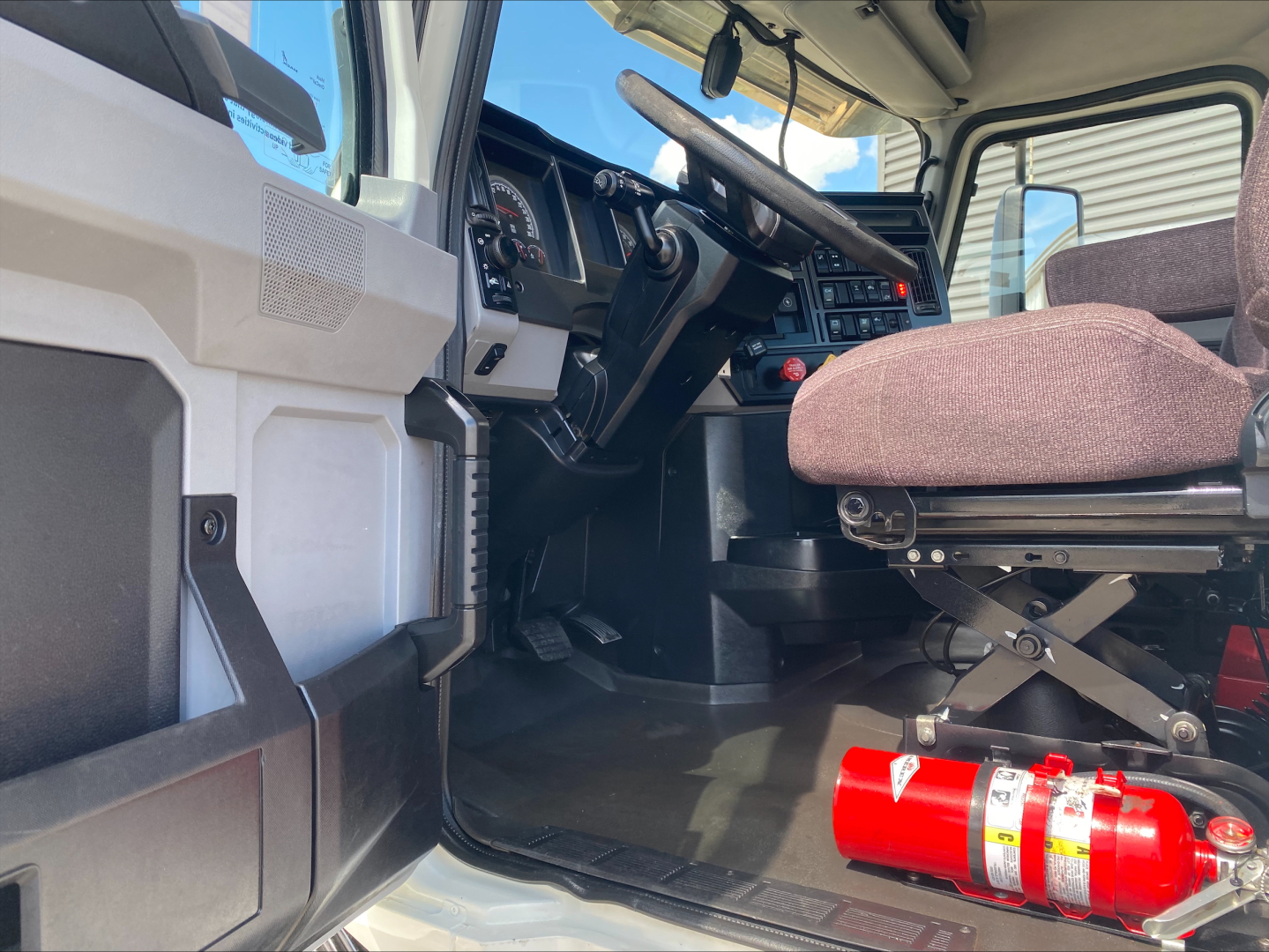 Cab interior of a 2021 Mack AN64T truck showing driver seat, steering wheel, dashboard, and a red fire extinguisher mounted near the floor.