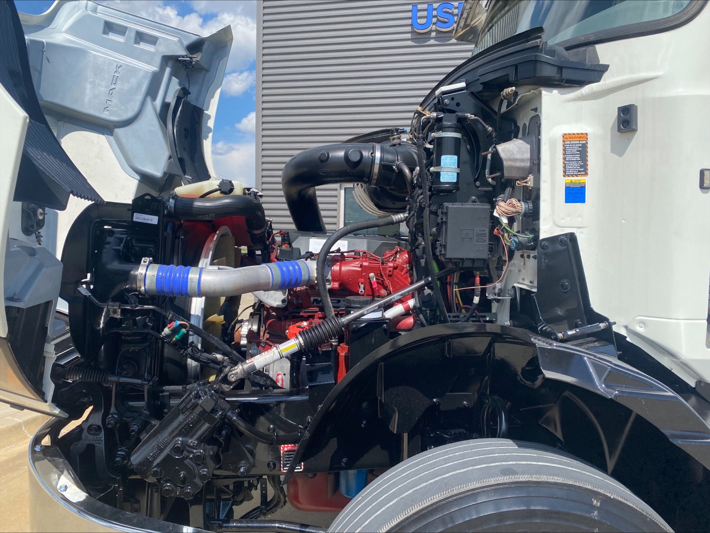 Engine bay of a 2021 Mack AN64T daycab showing red engine block, blue silicone hoses, black intake manifold, wiring, and exposed components.