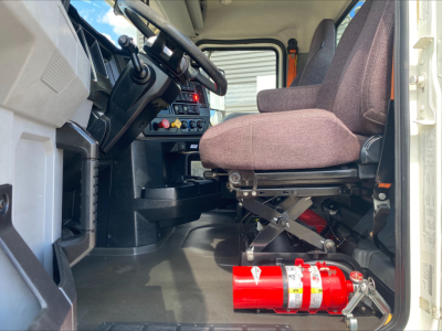 Cab interior of a 2021 Mack AN64T conventional daycab showing driver seat, steering column, dashboard controls, and a red fire extinguisher on the floor near the pedals.