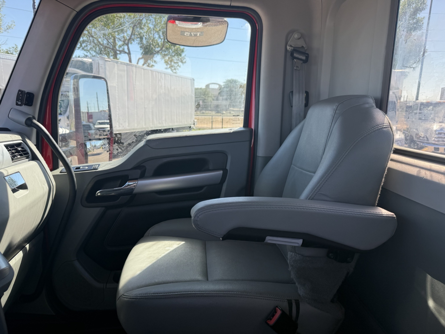 Truck cab interior showing driver seat and armrest from Kenworth T880 cab; front door panel and dashboard visible; light gray seats, window with exterior trailers.