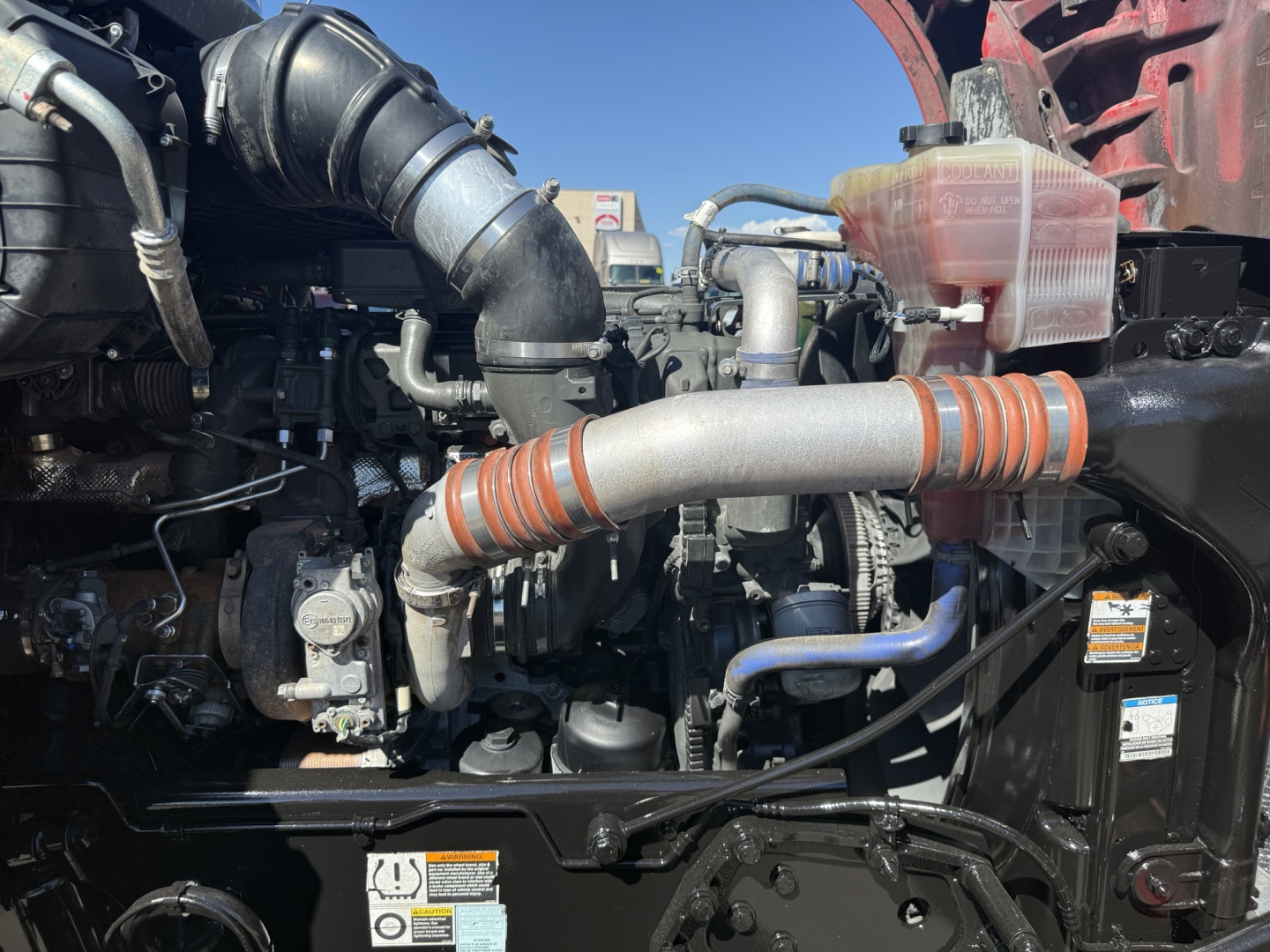 Kenworth T880 engine bay with intake tubing, coolant reservoir, and various hoses visible; orange heat shield bands around pipes.