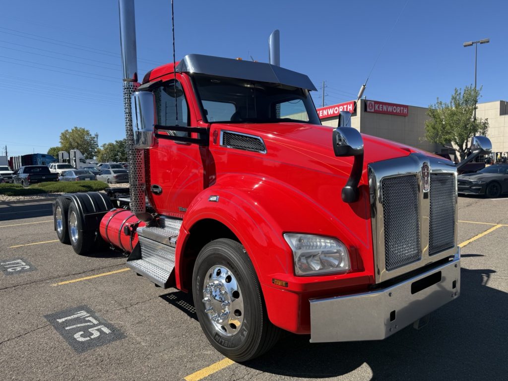 Kenworth T880 daytime cab truck in red, front-right view, chrome grille, dual exhaust stacks, chrome bumper, Kenworth dealership in background.