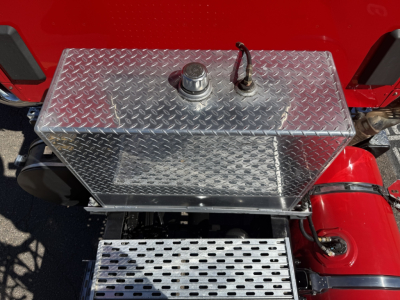 Aluminum diamond-plate fuel tank with filler cap and vent on a red Kenworth truck, visible under the cab with perforated aluminum step below.
