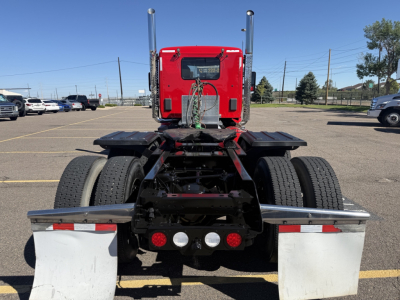 Kenworth T880 chassis rear view showing fifth wheel, dual rear axles, and mud flaps with chrome bumper; red cab in background.