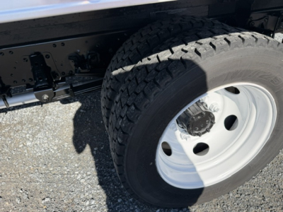 Isuzu NPR truck wheel and suspension components in view; solid white rim, dual rear wheels, tire tread visible; chassis and frame above with black-painted metal.