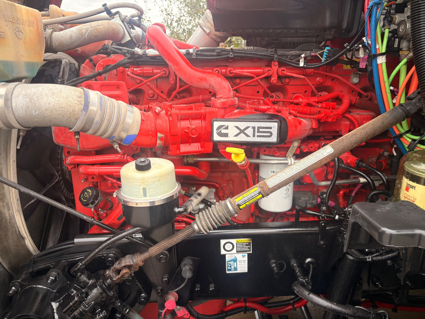 Kenworth CX15 engine bay showing bright red engine with CX15 badge and various hoses, tanks, and cables visible.