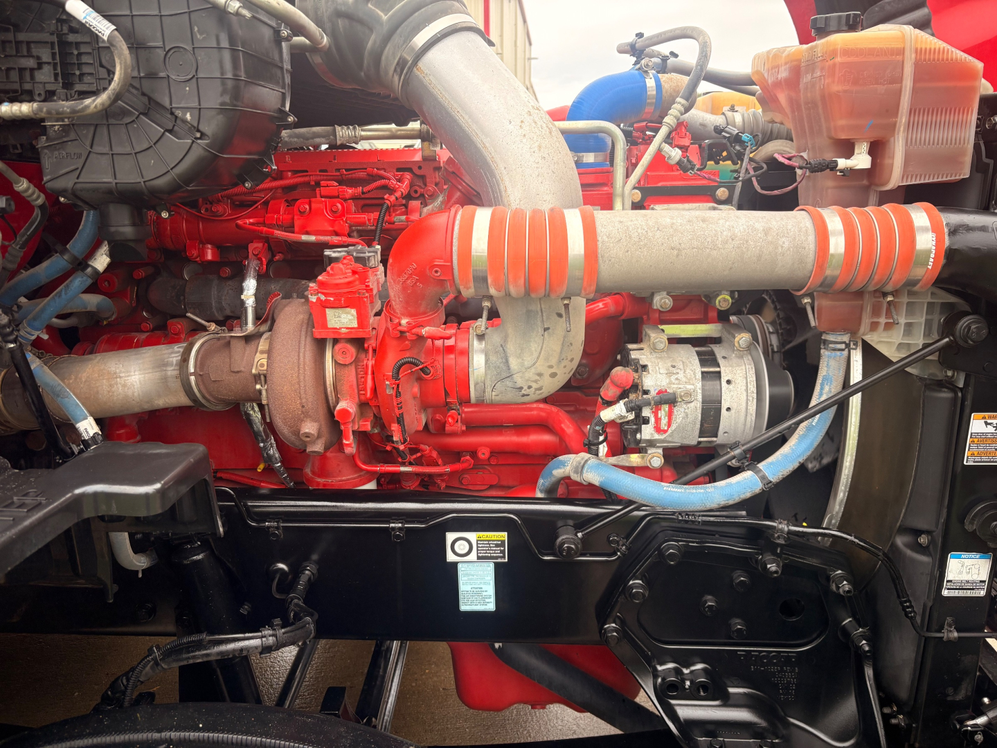 Kenworth T680 engine bay showing red engine block, intake piping, and coolant reservoir with orange hoses.