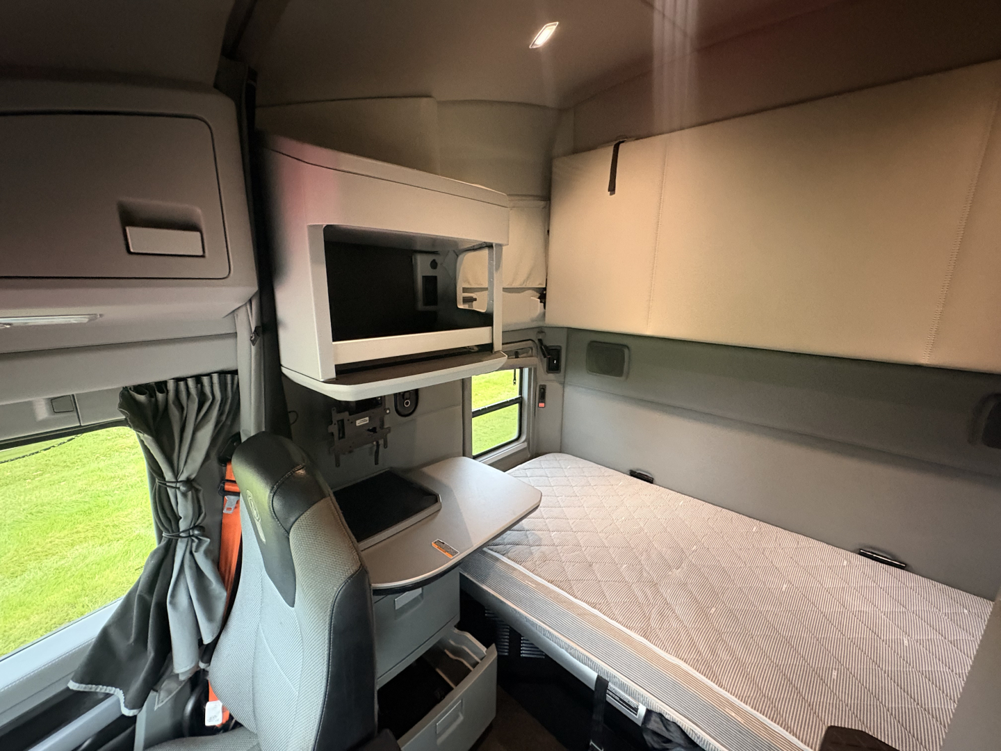 Kenworth T680 sleeper cab interior showing side bunk with mattress, overhead storage, and window; compact microwave shelf and curtain visible.