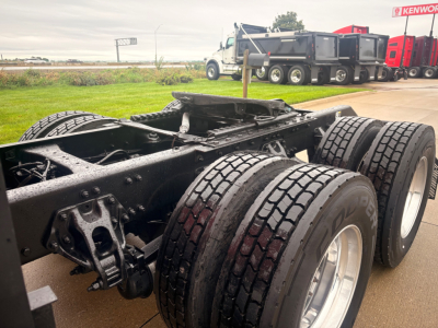 Kenworth truck chassis with dual rear axles and tires, fuel tank visible, trailer in background.