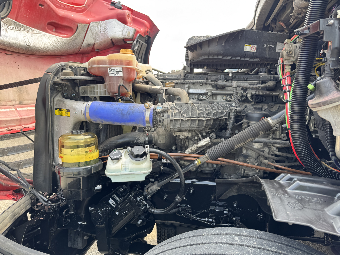 Freightliner Cascadia engine bay with visible blue intercooler hose, yellow coolant overflow bottle, black engine components, and orange hood area.