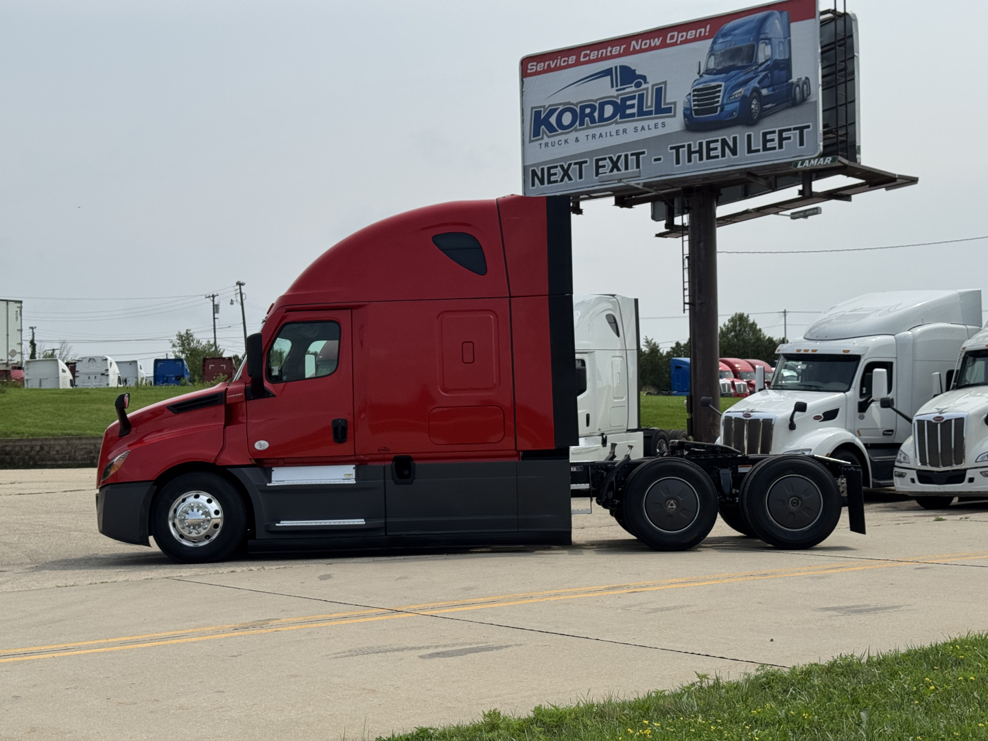 Truck tractor side view of a red Freightliner Cascadia 126 with sleeper cab, fuel tanks, and fifth wheel in a dealership lot.