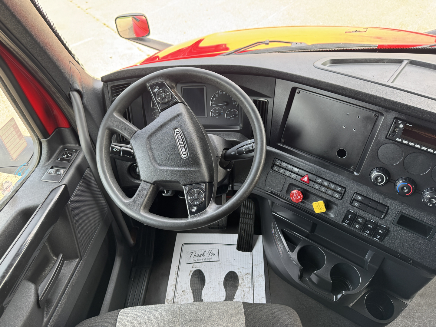 Cab interior of a Freightliner Cascadia truck showing steering wheel, dashboard, and center console with control panels and cup holders