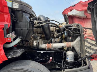 Truck engine bay of a 2023 Freightliner Cascadia 126 (conventional sleeper) showing engine, air intake, hoses, and coolant reservoir. Visible components include radiator, intercooler piping, oil filter, and top of wheel well.