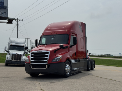 2013 Freightliner Cascadia 126-style sleeper cab truck in red with chrome grille, parked on a lot with other trucks in background.