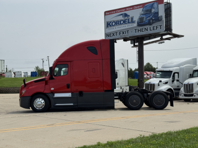 Truck tractor side view of a red Freightliner Cascadia 126 with sleeper cab, fuel tanks, and fifth wheel in a dealership lot.