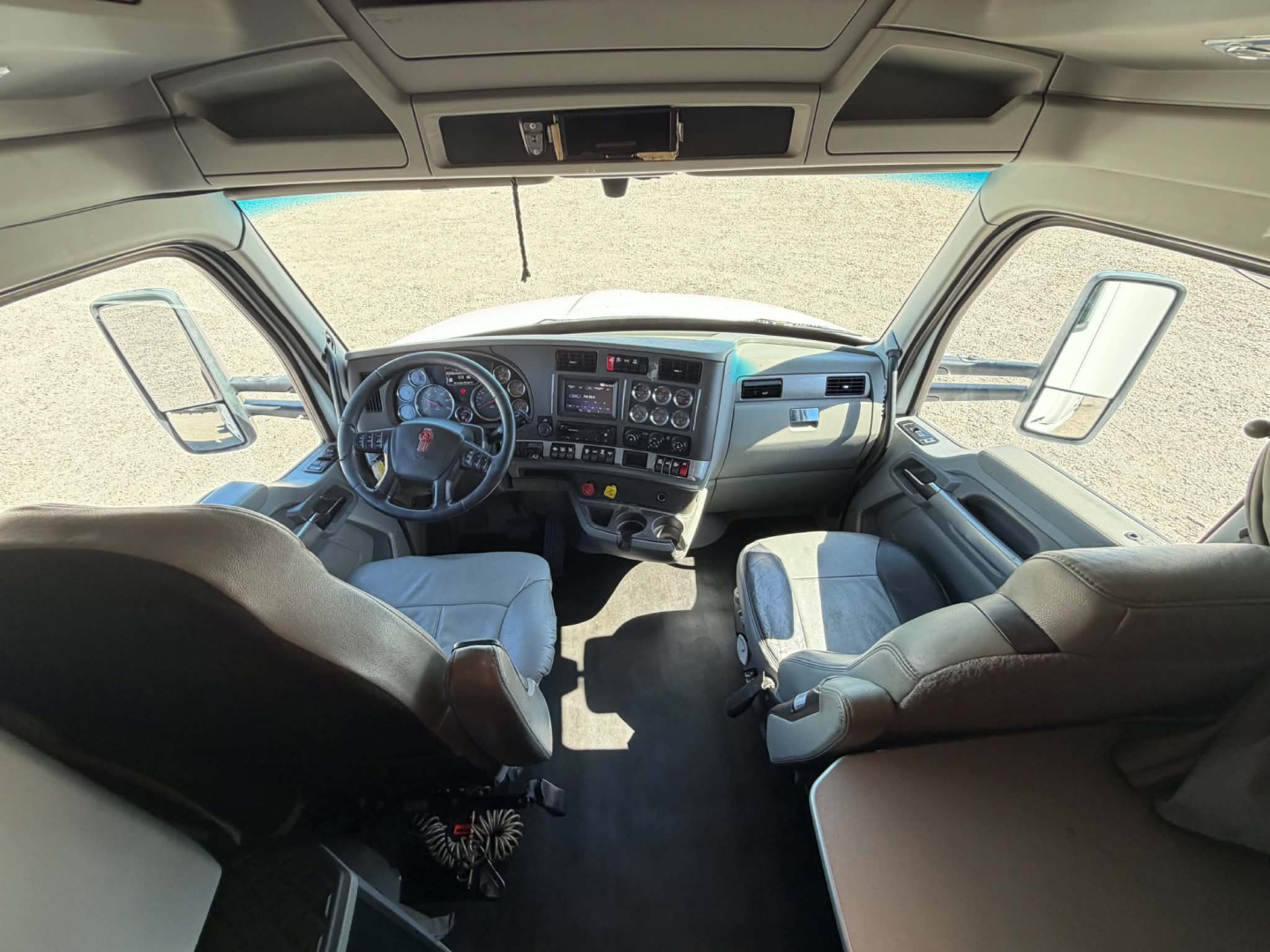 Cab interior of a 2019 Kenworth T680 tractor from front passenger seat perspective, showing steering wheel, dashboard with gauges, center console, sun visors, and two Adjusted driver and passenger seats.