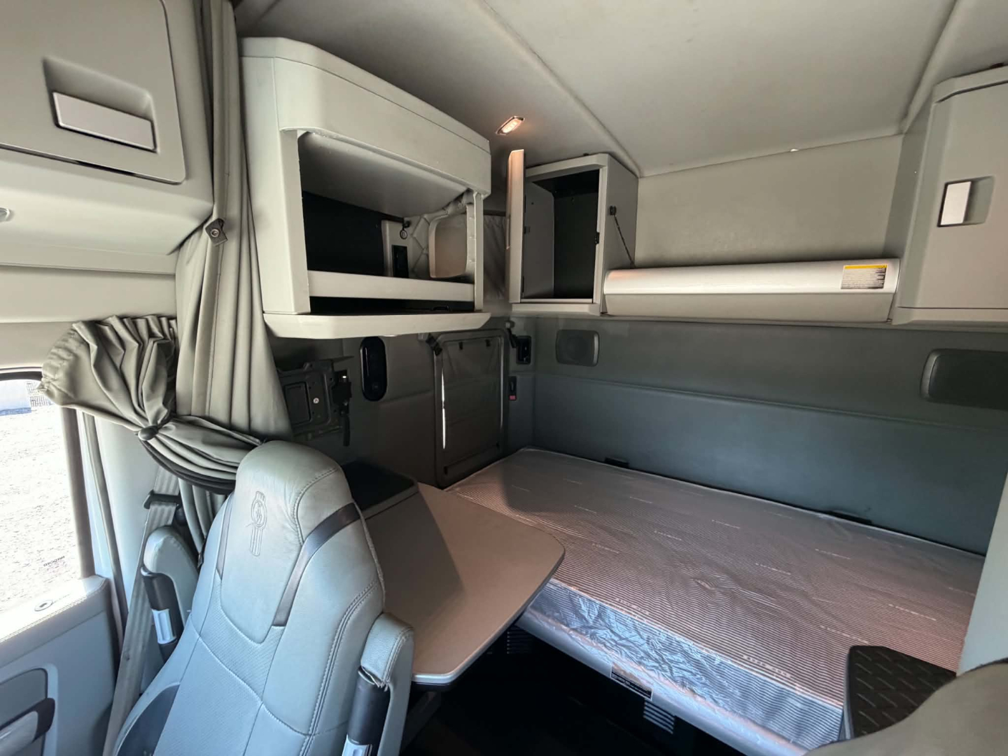 Interior cab view of a 2019 Kenworth T680 sleeper: bunk bed, overhead cabinets, window curtains, and workspace table visible.