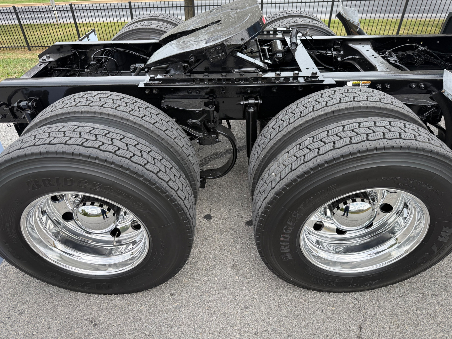 Kenworth truck chassis with dual rear axles and large tires; no cab, showing exposed frame, suspension, and chrome wheels.