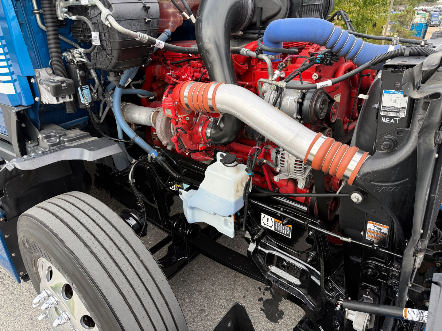 Kenworth T680 engine bay showing red engine block with orange hose clamps, blue coolant hose, white coolant reservoir, and black radiator supports.