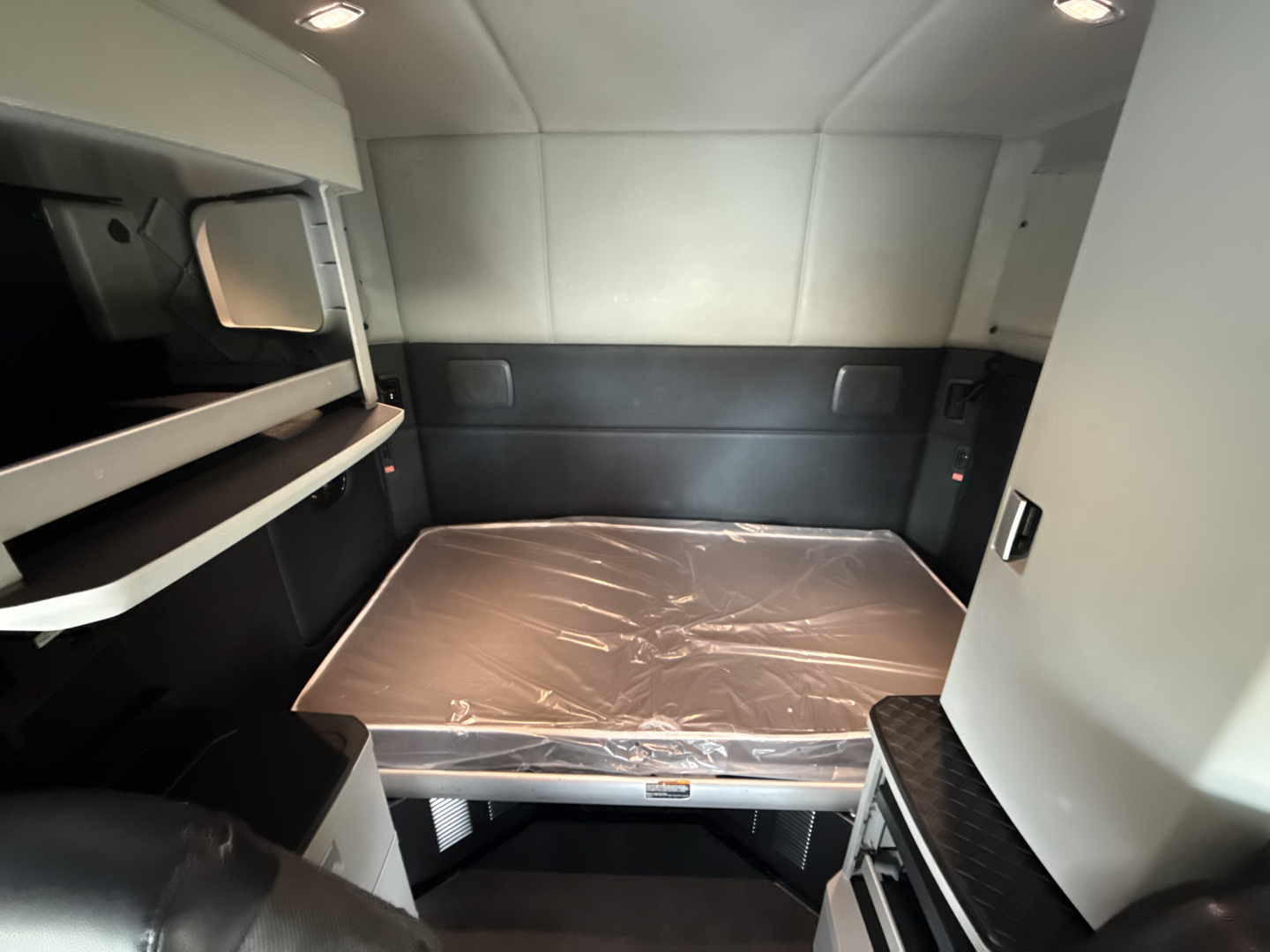 Kenworth T680 sleeper cab interior showing the bed on a plastic-covered mattress, side cabinets, and upper bunk shelf in a compact sleeper area.