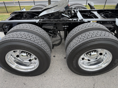 Kenworth truck chassis with dual rear axles and large tires; no cab, showing exposed frame, suspension, and chrome wheels.