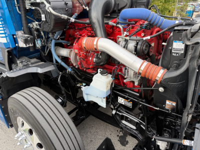 Kenworth T680 engine bay showing red engine block with orange hose clamps, blue coolant hose, white coolant reservoir, and black radiator supports.