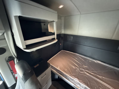 Kenworth T680 sleeper cab interior showing side bunk with plastic-wrapped bed, overhead storage/ashtray area, small shelves, and plastic-wrapped seat; cabin lighting visible.
