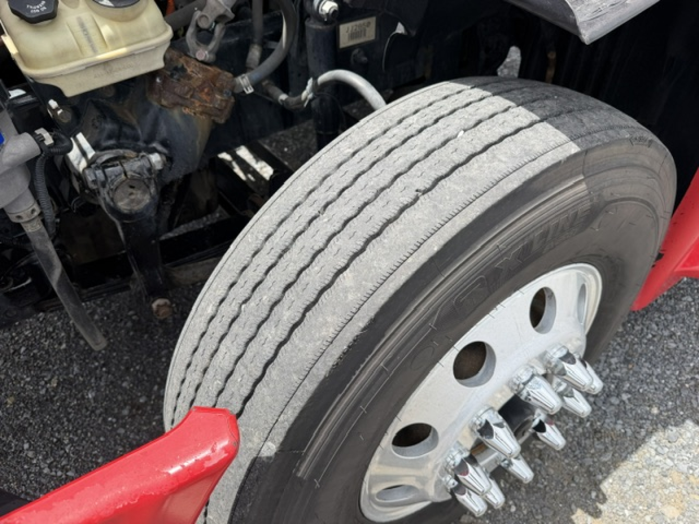 Truck wheel and front axle area of a Freightliner Cascadia CA125DC showing large dual rear tires, front wheel with multi-lug pattern and surrounding engine bay components.