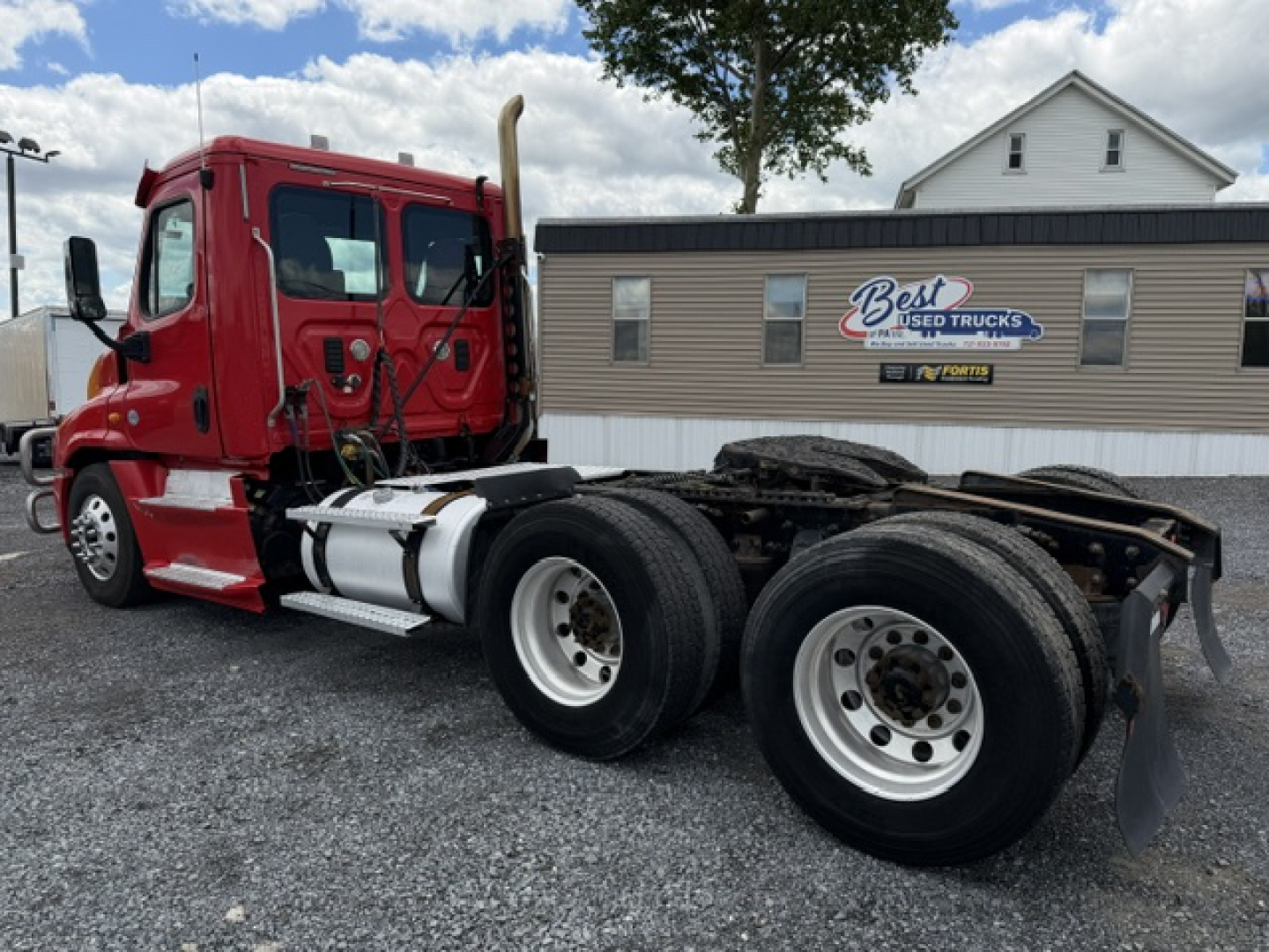Red Freightliner Cascadia CA125DC tractor side view showing dual rear axles and chassis. Background includes building with Best Used Trucks sign and cloudy sky.