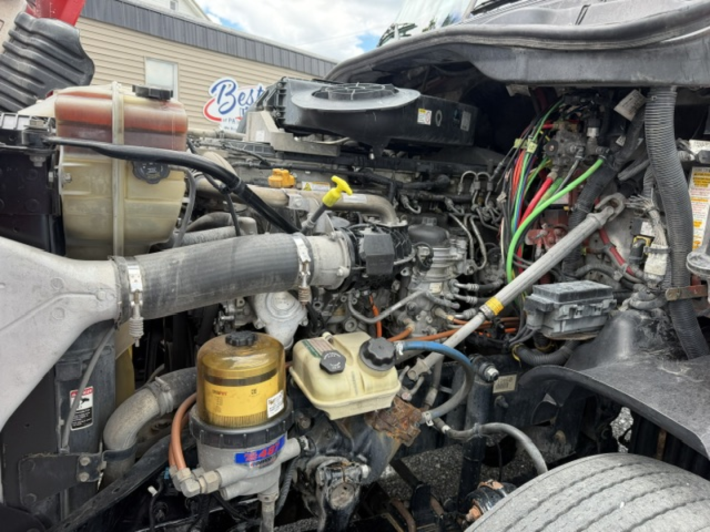 Truck engine bay view showing various hoses, wiring, and fluid reservoirs in a Freightliner Cascadia engine compartment.
