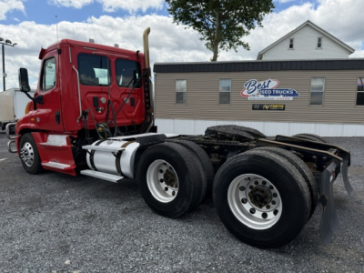 Red Freightliner Cascadia CA125DC tractor side view showing dual rear axles and chassis. Background includes building with Best Used Trucks sign and cloudy sky.