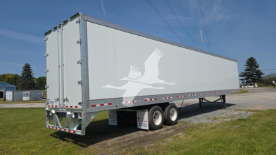 Trailer side view showing a large gray Vanguard VXP high base rail trailer with white crane-like graphic on the side and dual rear doors.