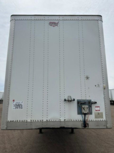 Rear view of a metal trailer with riveted panels and a small locking mechanism near the bottom right; stickers and a faded logo on the upper center.