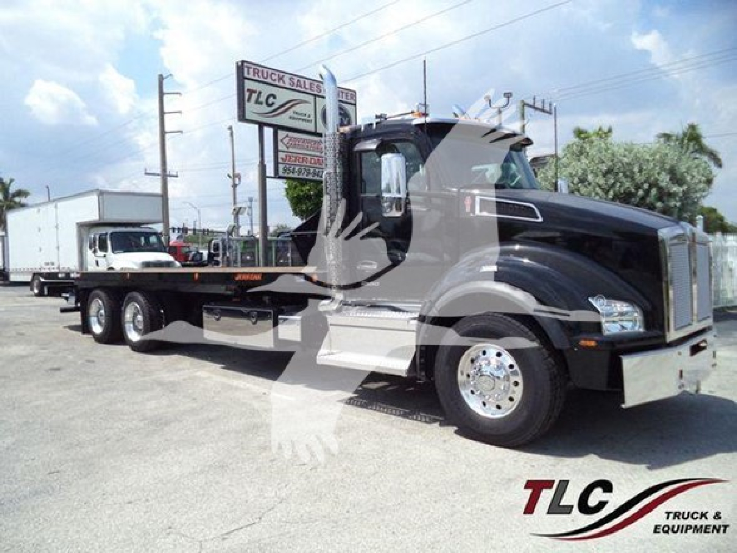 Kenworth T880 rollback truck in a dealership lot, black cab with chrome front bumper and flatbed; TLC Truck Sales sign visible in background.