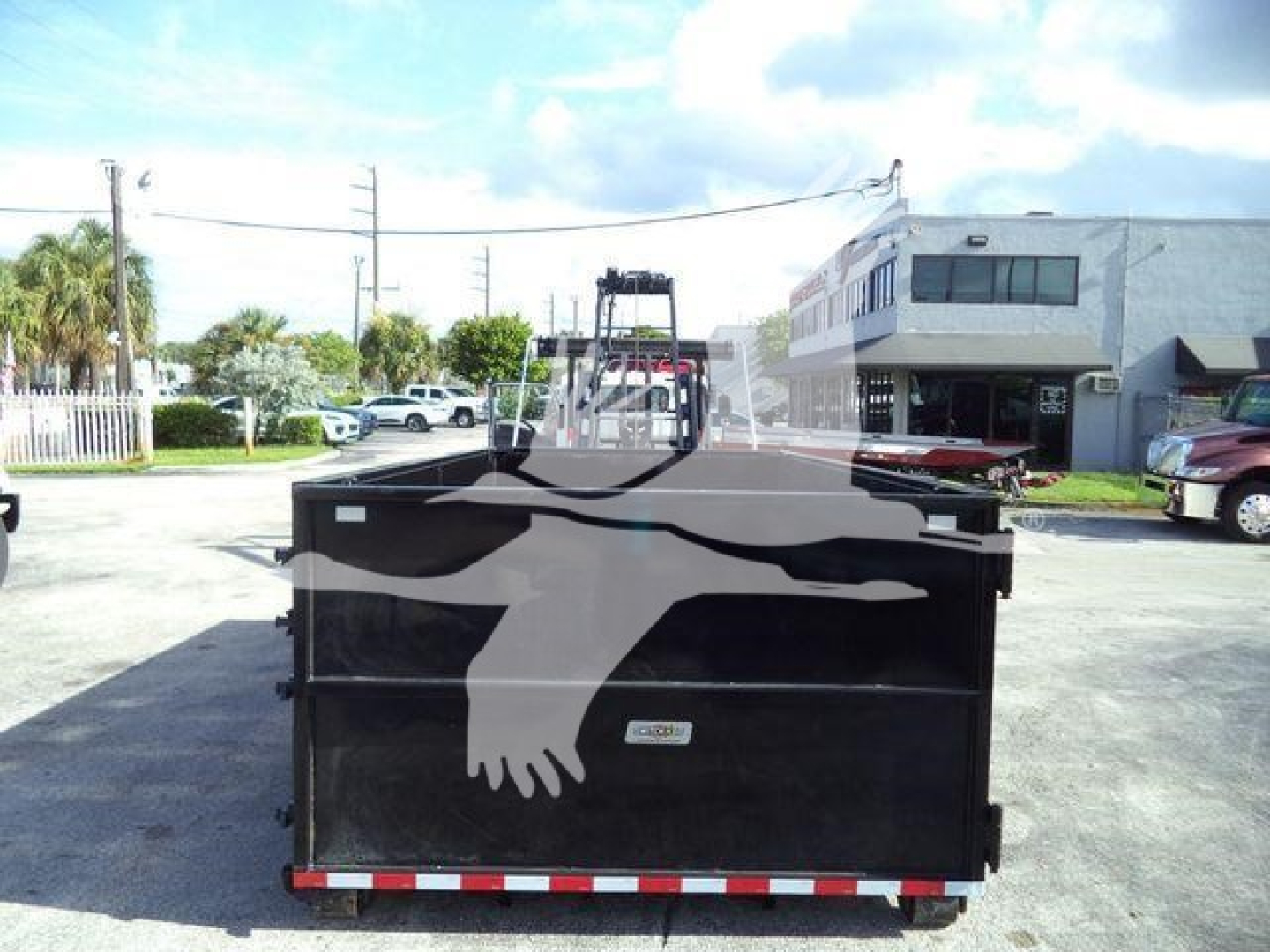 Isuzu NPRR roll-off truck bed with rear view and faded logo on black dumpster in foreground; urban lot and building in background.