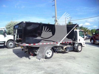 Isuzu NPR/NRR roll-off truck in a lot, black dump body, white cab, side view showing rear wheel, hydraulic lines, and trash container. Visible text on side includes vendor tags and markings but no legible model numbers.
