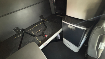 Kenworth T680 sleeper cab interior storage area with seat and small fridge unit, visible wiring and rails.