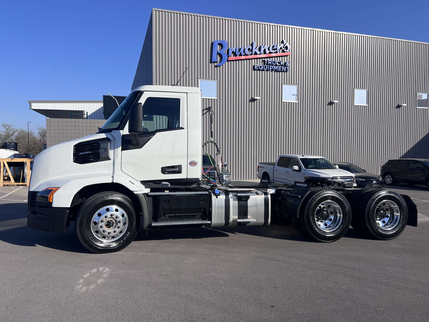 Isometric view of a white 2027 Mack AN64T conventional daycab on a dealership lot with a Bracknek Equipment building in background.