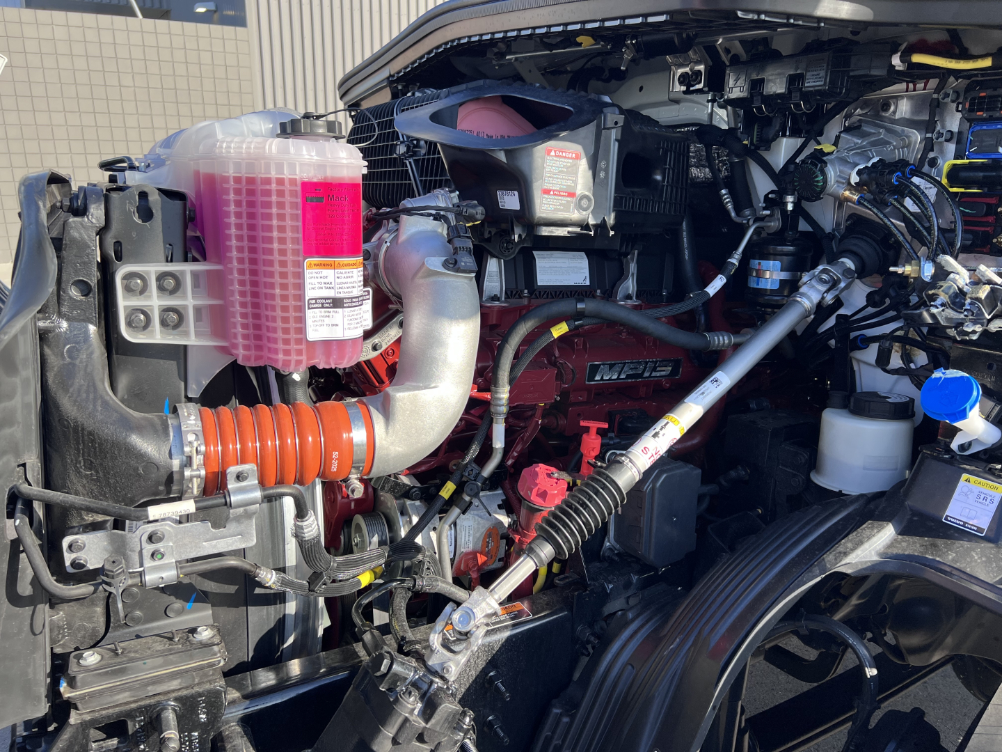 Front engine bay of a 2027 Mack AN64T Daycab showing coolant reservoir, pink coolant/antifreeze, pink fluid container, hoses, belts, and engine components.