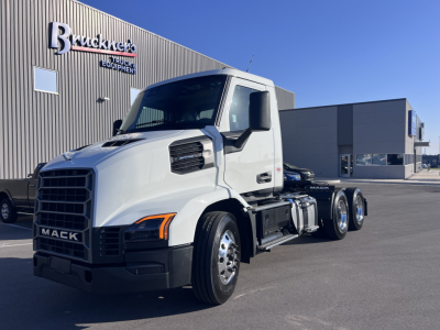 Mack AN64T conventional daycab tractor in white, front-left angle showing chrome grille, large side mirrors, and dual rear axles; Buckner’s equipment lot in background with warehouse.