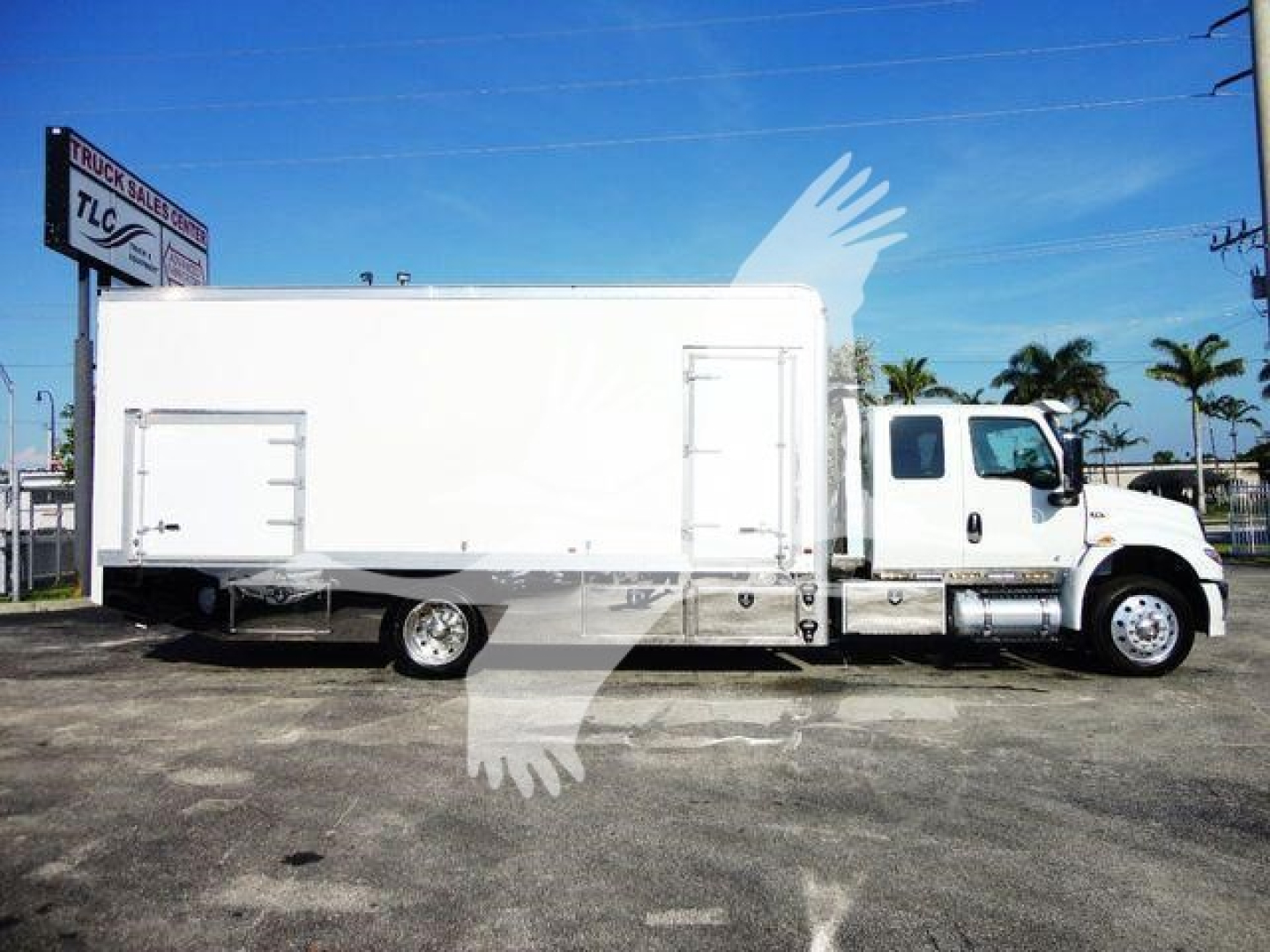 Truck side view of a 2022 International MV rollback/box truck, with a large white cargo box, cab at right, wheels visible, palm trees and dealership sign in background.