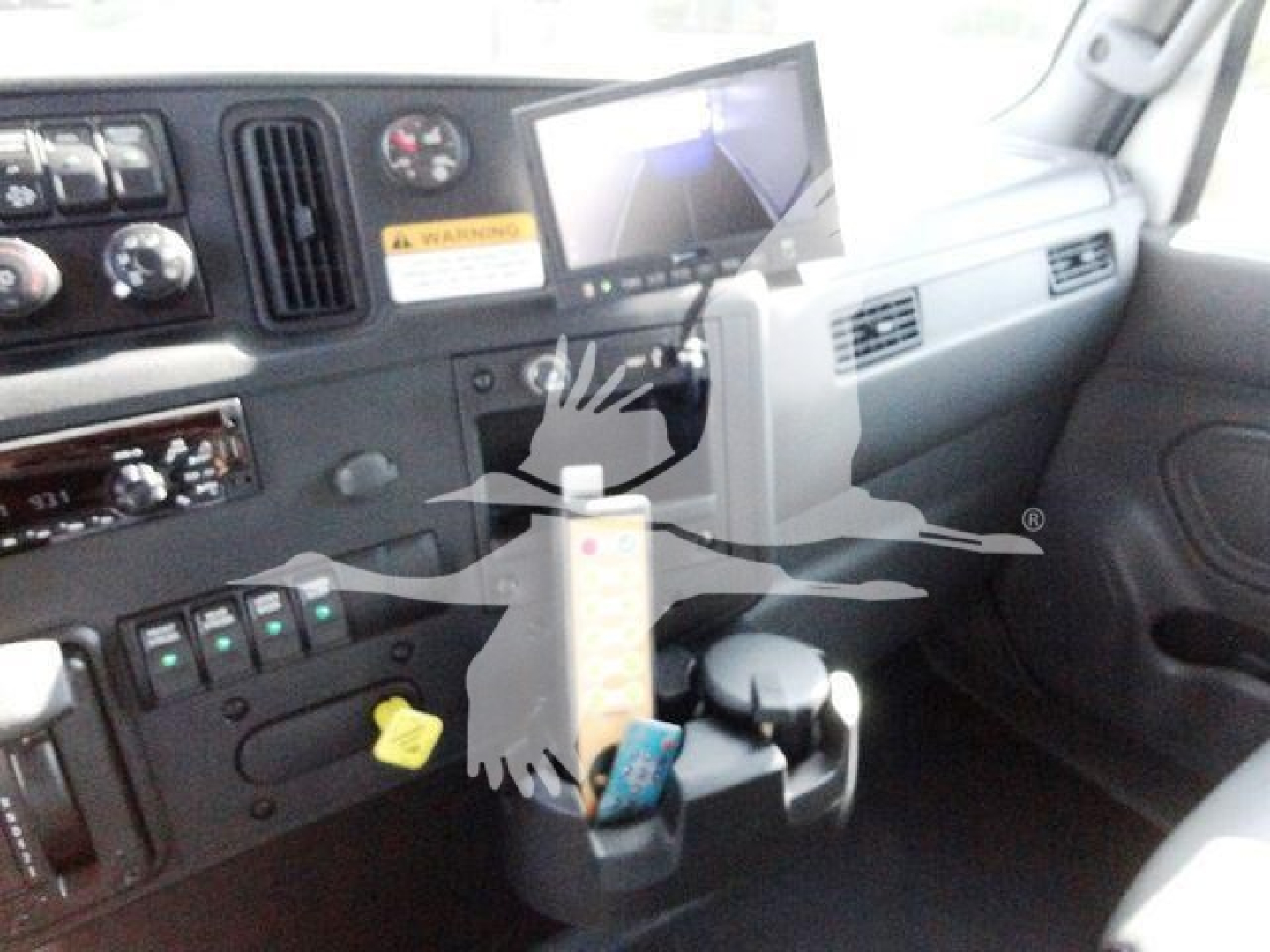 Dashboard and steering wheel area of a truck cab showing instrument cluster, control switches, a hanging card, and a cup holder with a blue object.