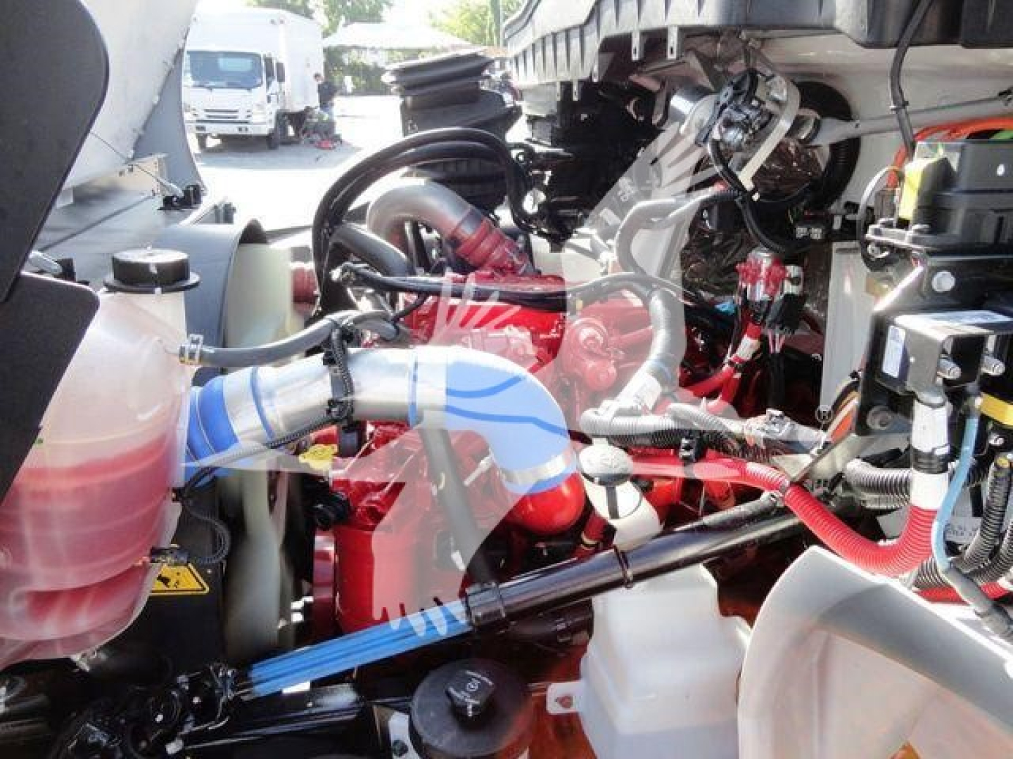Truck engine bay with red engine, hoses, and blue coolant hose visible; various reservoirs and components in a MV International truck setup.