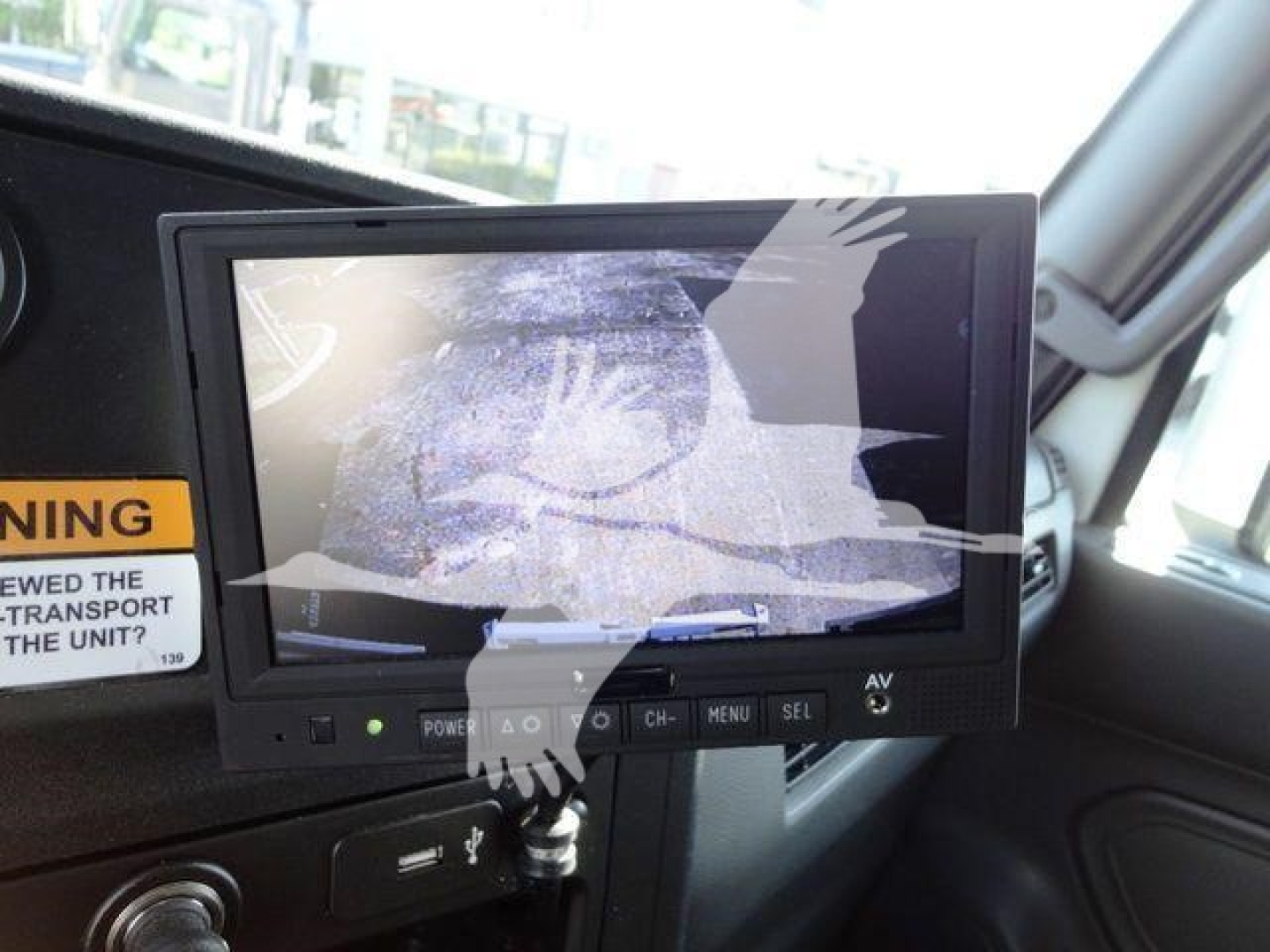 Cab interior showing a monitor/display mounted on the dashboard with a protective screen; visible control buttons below; power and AV indicators; part of the truck interior.