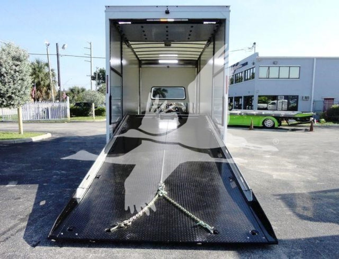 Truck cargo area with a lowered ramp on a flatbed trailer, interior walls white, cab entrance visible at far end, chain hooks on ramp edges.