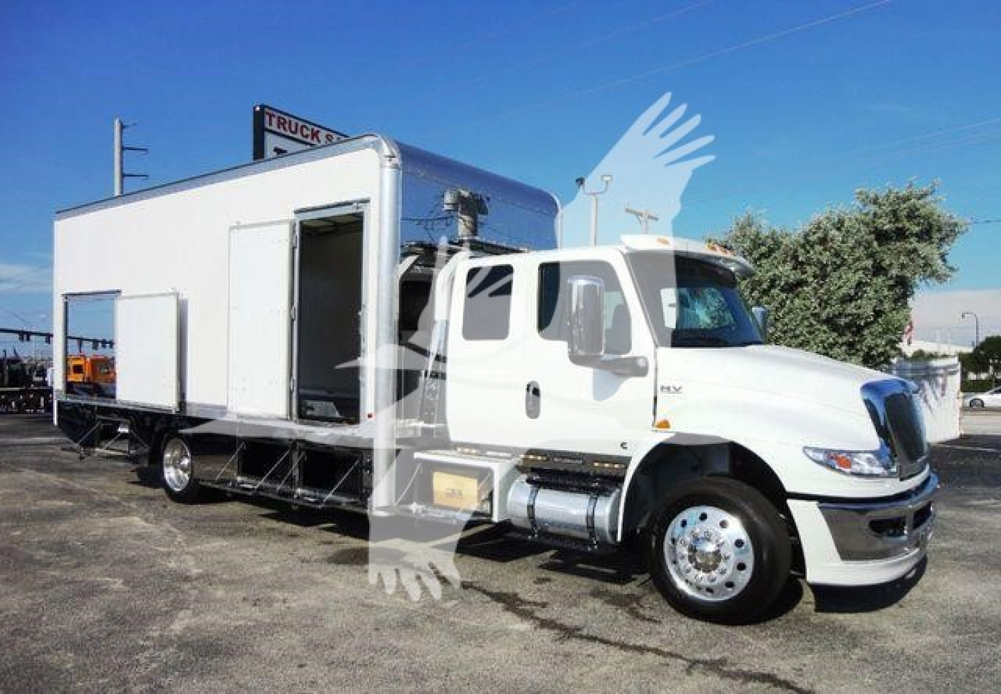 Truck mounted rollback wrecker body on a white International MV cab chassis. Side door open, living quarters module behind cab, logo on front grille visible.