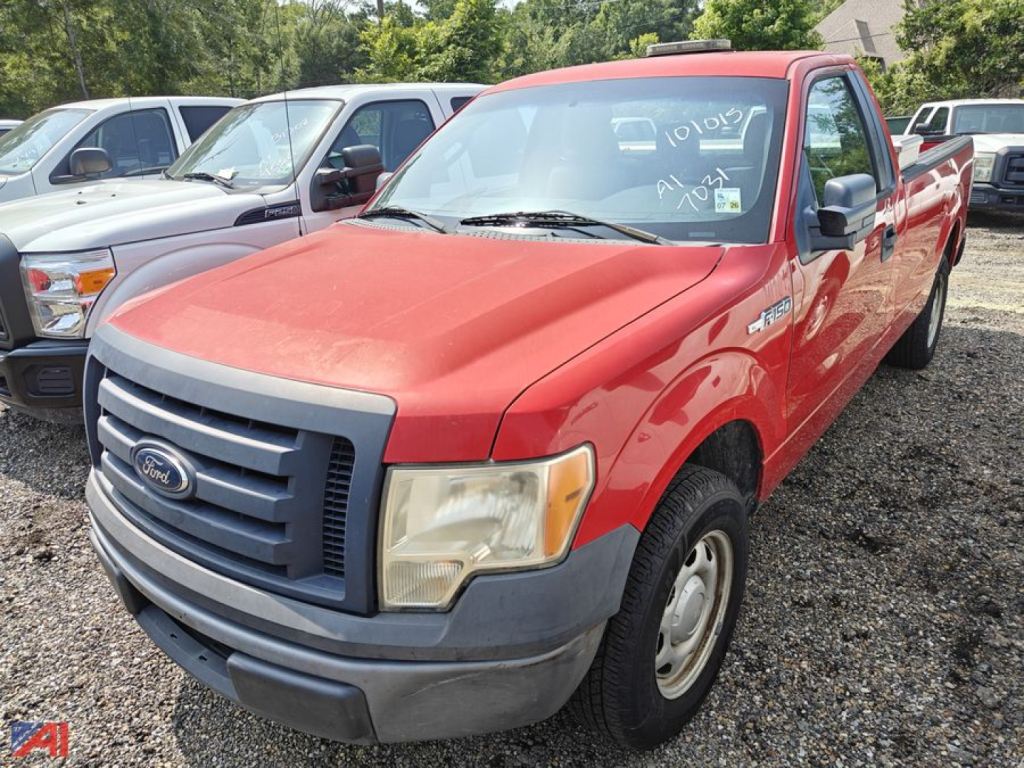 Red 2011 Ford F-150 pickup truck in a lot, front three-quarter view showing Ford grille and bumper. Windshield has white handwritten markings. Other vehicles in background.